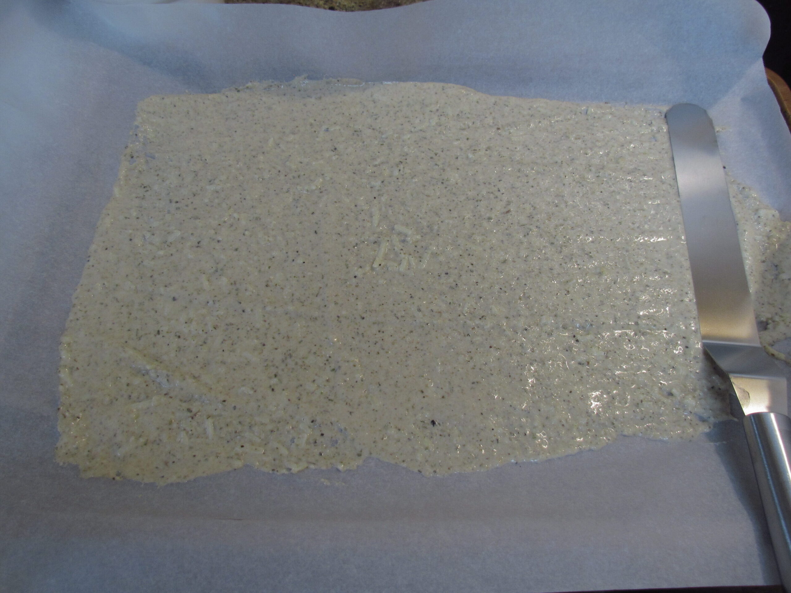 picture of sourdough cracker batter spread out on parchment lined pan with a spreading spatula