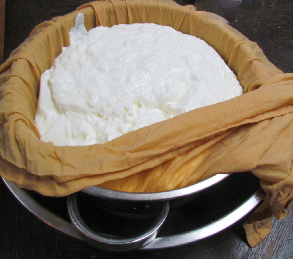 Picture of yogurt inside a tea towel inside a strainer inside a bowl.